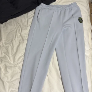 Ljusblå mjukisbyxor från Lacoste - Snygga ljusblå mjukisbyxor från Lacoste med regular fit och elastisk midja. Byxorna har raka ben, sidofickor och ett broderat Lacoste-märke på höften. Materialet är mjukt och strukturerat. Storlek S men passar M