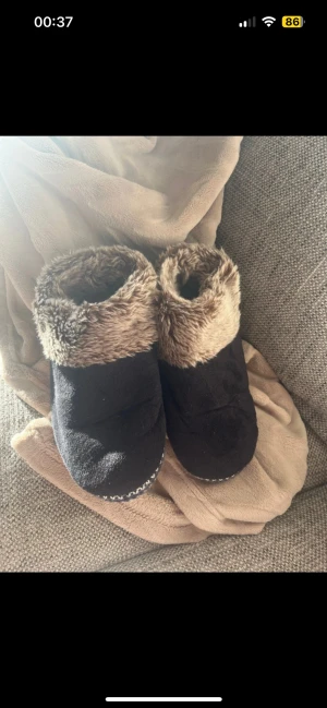 Svarta tofflor med fuskpäls - Mysiga svarta tofflor med mjuk fuskpäls i brunt upptill och prickig sula. Utsidan är i mockaliknande material. Perfekta för kalla dagar hemma.