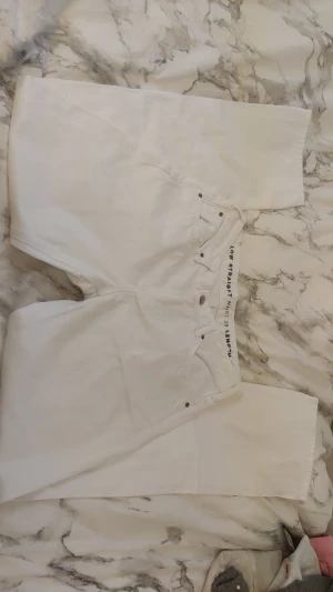 Vita straight jeans från Never Denim - Säljer ett par vita jeans från Never Denim i modellen Low Straight. Byxorna har låg midja, raka ben och klassisk femficksdesign. Tillverkade i bomullstyg med snygga detaljer i metall och läderpatch bak. Perfekta för en clean och fräsch look.