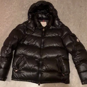 Svart Moncler pufferjacka med huva - Säljer en svart pufferjacka från Moncler med huva och dragkedja framtill. Jackan har två sidofickor med dragkedja och Moncler-logga på ärmen. Den är dunfylld och har en glansig finish som ger en cool look. Perfekt för kalla dagar.