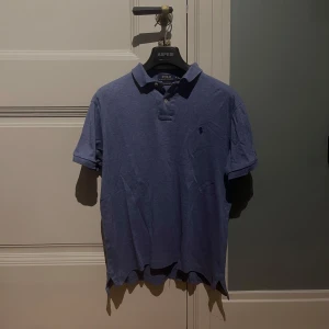 Blå pikétröja från Polo Ralph Lauren - Säljer en blå pikétröja från Polo Ralph Lauren i custom slim fit. Tröjan har klassisk krage, två knappar och korta ärmar. Materialet är mjuk bomull och den har den ikoniska lilla loggan broderad på bröstet. Perfekt för dig som gillar stilren och enkel design.
