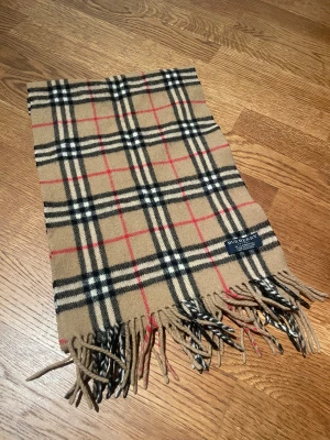 Halsduk från Burberry i ull - Klassisk halsduk från Burberry i beige, svart, vitt och rött med ikoniskt rutmönster. Tillverkad i 100% lammull och har fransar i ändarna. Perfekt accessoar för att lyfta din outfit med en tidlös brittisk touch.
