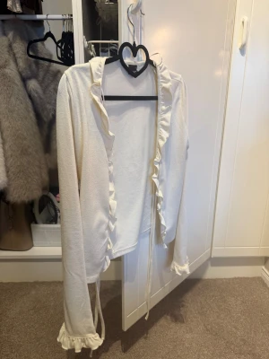 Vit omlottopp från Gina Tricot - Säljer en söt vit omlottopp från Gina Tricot i storlek S. Toppen har långa ärmar, volangdetaljer längs kanterna och knytband framtill. Materialet är mjukt och känns som bomull eller polyester. 