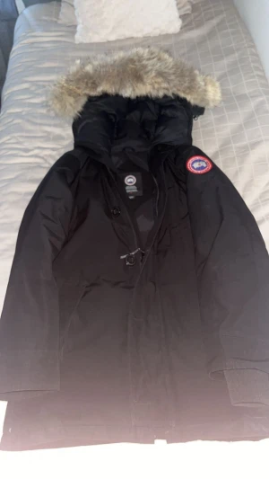 Svart parkas från Canada Goose - Svart parkas från Canada Goose med klassisk logga på ärmen och stor huva med fluffig päls. Jackan har dragkedja och knappar framtill samt snörning vid midjan. Perfekt för kalla dagar med sin långa modell och varma material funkaf både för s och m.