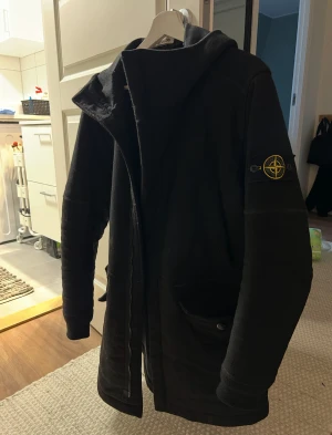 Svart rock från Stone Island - Svart parkajacka/rock från Stone Island med huva och klassisk logotyp på ärmen. Jackan är tillverkad i stretchigt ullmaterial med flera lager för extra komfort och vindmotstånd. Den har dragkedja framtill och praktiska fickor med knappstängning.