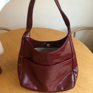 Vinröd axelväska  - Vinröd axelväska i glansigt faux leather.   Väskan är rymlig och rymmer en 13-tums MacBook utan problem. Den stängs med magnetknapp (ingen dragkedja).   Insidan har ett separat innerfack med dragkedja. På utsidan finns dessutom ett ytterfack.  En perfekt vardagsväska som funkar till skola, jobb eller ärenden.  Köpt på TikTok Shop och knappt använd. Har en svag faux leather-doft, men inget störande.