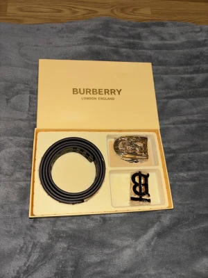 Burberry bälte med två spännen - Säljer ett Burberry bälte i klassisk design med två utbytbara spännen – ett i guld med Burberry-logga och ett svart TB-monogram. Ena sidan är rutigt i beige, svart och rött, andra sidan är helsvart. Bältet levereras i originalask.pris kan diskuteras vid ett snabbt köp