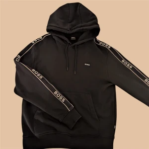 Svart hoodie från BOSS med logga - Svart hoodie från BOSS med huva och snörning. Snygga vita BOSS-loggor längs båda ärmarna och en liten logga på bröstet. Mjuk insida och ribbade muddar. Perfekt för dig som gillar streetwear och vill ha en clean look.