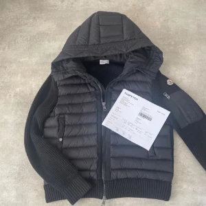 Svart Moncler cardigan med huva - Svart pufferjacka från Moncler med stickade ärmar och quiltad kropp. Jackan har en stor huva, dragkedja framtill och Moncler-logga på ärmen. Snygg ribbad kant vid ärmslut och nederkant, samt praktisk ficka med dragkedja på ärmen.