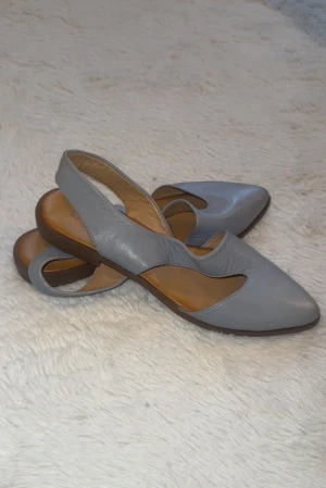 Ljusblåa slingback sandaler från Bueno - Snygga ljusblåa sandaler från Bueno med slingback och spetsig tå. Skorna är tillverkade i mjukt skinn med en unik utskuren design på ovansidan och har en platt brun sula. Perfekt för dig som gillar stilrena och moderna detaljer.