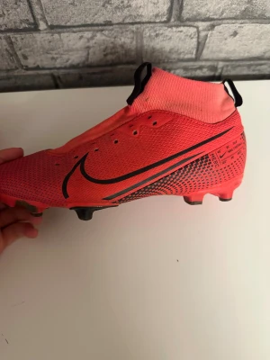 Röda nike mercurial fotbollsskor  - Säljer ett par röda Nike fotbollsskor med svart swoosh och strumpkrage för extra stöd runt ankeln. Skorna har platt sula med dobbar för gräsplan och är tillverkade i syntetmaterial med ventilerande mesh. Snygg design med svarta detaljer och mönster på sidorna.