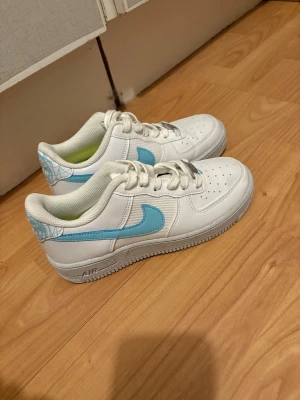 Nike Air Force 1 vit/blå sneakers - Nike Air Force 1 sneakers i vitt skinn med ljusblå swoosh och grafiskt mönster på hälen. Klassisk låg modell med rund tå, snörning och platt sula. Perfekt för dig som gillar stilrena och fräscha färgkombinationer. Endast använda en gång pga att dom var för små.