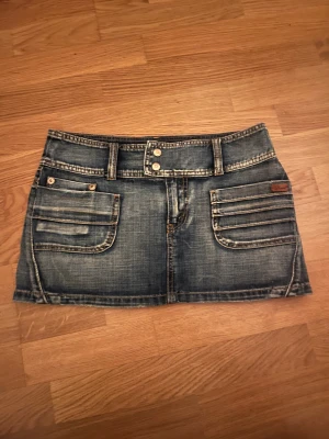 PHARD jeanskjol med fickor - Säljer en cool kort jeanskjol från PHARD. Kjolen har stora fickor både fram och bak samt ett brunt lädermärke med logga bak. Den är i klassisk blå denim med slitna detaljer och kontrastsömmar. Kjolen är i storlek 27 men jag skulle säga att den passar xs-s💕🥰⭐️