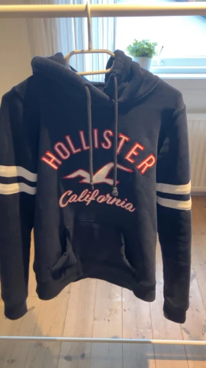 Mörkblå Hollister hoodie med tryck - Snygg mörkblå hoodie från Hollister i storlek s/xs med stort rött och vitt tryck på bröstet och texten 'California'. Tröjan har huva med snörning, vita ränder på ärmarna och en klassisk känguruficka framtill. Perfekt för chill dagar och streetstyle.