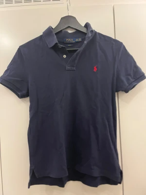 Mörkblå Polo Ralph Lauren piké - Snygg mörkblå pikétröja från Polo Ralph Lauren med klassisk krage och två knappar. Slim fit-modell med korta ärmar och det ikoniska röda broderade polospelaren på bröstet. Tillverkad i mjuk bomull som är skön mot huden.