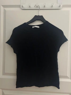 Svart topp från Zara - Svart topp från Zara med korta ärmar och rund hals. Materialet är stickat och har en lätt transparent känsla, vilket ger en cool vibe. Perfekt att styla med jeans eller kjol för en enkel men snygg look.