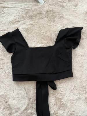 Svart croppad topp - Säljer en svart croppad topp med korta volangärmar och snygg fyrkantig urringning. Toppen har ett brett band som knyts i ryggen. Helt oanvänd och är i storlek Xs