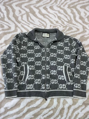 Grå kofta med GG-mönster från Gucci - Säljer en grå stickad kofta från Gucci med ikoniskt vitt GG-mönster över hela tröjan. Koftan har dragkedja framtill, ribbad krage och muddar samt två fickor med vit kant. Perfekt för dig som gillar lyxiga detaljer och snygg design.