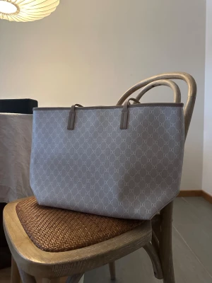 Beige shoppingväska från DKNY - Stilren shoppingväska från Don Donna/Glitter i beige med monogrammönster över hela väskan. Väskan har två rejäla handtag och är tillverkad i tålig syntet med detaljer i skinnimitation. Perfekt storlek för att få plats med allt du behöver.