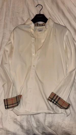 Burberry Sherfield Stretch Bomullspoplin Skjorta - Snygg vit skjorta från Burberry med klassiska rutiga detaljer på insidan av manschetterna. Skjortan har knappar framtill, lång ärm och broderad logga på bröstet. Perfekt för dig som gillar stilrena plagg med en twist. Nypris: 4950kr. Storlek: S. Kan fraktas. Tar endast Swish. Köp sker utanför denna appen, dvs ej med köparskydd.