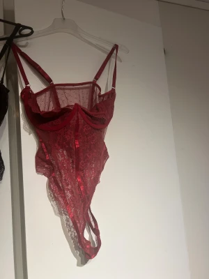 Röd spetskorsett med smala axelband - Säljer en röd korsett med smala axelband och genomskinlig spets. Modellen har markerade kupor och dekorativa band framtill. Spetsen har ett snyggt mönster och ger en feminin vibe. Perfekt för dig som vill ha något extra till din outfit.