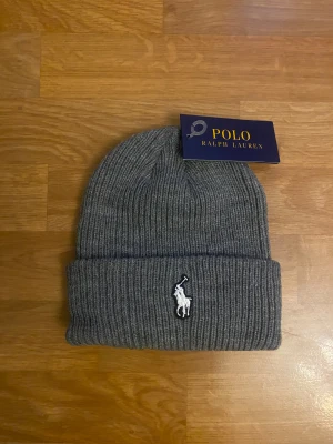 Polo Ralph Lauren Mössa - Snygg grå ribbstickad mössa från Polo Ralph Lauren med klassisk broderad logga i vitt framtill. Mössan har uppvikt kant och är tillverkad i mjukt material som håller dig varm och stilren under kyliga dagar.