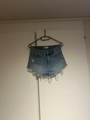 Blåa  jeansshorts med fransar - Säljer ett par blåa denimshorts från Gina Tricot med råa fransade kanter. Shortsen har slitna detaljer både fram och bak samt klassiska fem fickor. Perfekta för sommaren och ger en cool, avslappnad vibe.