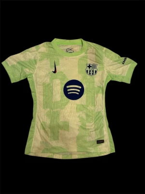 FC Barcelona Nike fotbollströja grön - Snygg FC Barcelona fotbollströja från Nike i ljusgrönt med unikt geometriskt mönster. Tröjan har klubbmärke, Nike-logga och Spotify-tryck på bröstet samt UNHCR-tryck på ryggen. Tillverkad i lätt och ventilerande funktionsmaterial, perfekt för match eller träning.