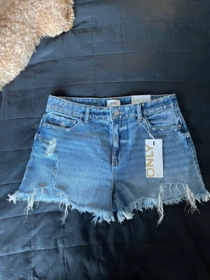 Blåa high waist jeansshorts från ONLY - Snygga blå jeansshorts från ONLY med hög midja, slitna detaljer och fransig kant. Shortsen har nitar längs midjan och fickorna, klassisk femficksmodell och är perfekta för en chill sommarlook. Aldrig använda så nyskick☺️😍💕De är ganska små i storleken så de passar också strl M😁😇