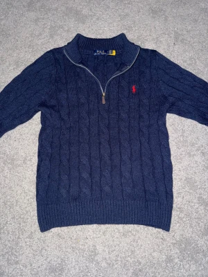 Marinblå stickad half zip från Polo Ralph Lauren - Snygg marinblå stickad tröja från Polo Ralph Lauren med half zip och kabelstickat mönster. Tröjan har långärmad design, ribbad krage och muddar samt röd broderad logga på bröstet. Perfekt för dig som gillar klassisk stil med en modern touch.