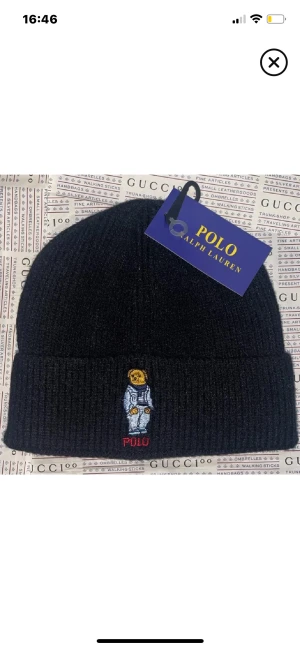 Svart ribbad mössa Polo Ralph Lauren - Svart ribbstickad mössa från Polo Ralph Lauren med broderad Polo-björn i astronautdräkt framtill. Klassisk uppvikt kant och mjukt material som håller dig varm. Perfekt accessoar för att lyfta din streetstyle.