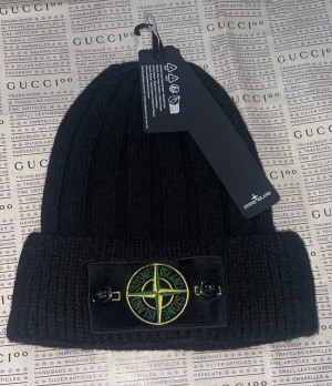 Svart ribbad mössa från Stone Island - Svart ribbstickad mössa från Stone Island med klassisk logopatch framtill i grönt och gult. Mössan har uppvikt kant och är tillverkad i mjuk ull, perfekt för kalla dagar. Snygg och enkel att matcha med din streetwear eller vinteroutfit.