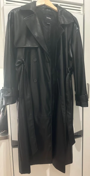 Tenchcoat från Pull&Bear - Svart trenchcoat från Pull&Bear i snyggt syntetskinn med klassisk krage, bälte i midjan och knappar framtill. Jackan har långa ärmar med detaljer vid mudden och en stilren, rak passform som ger en cool vibe. Perfekt för dig som gillar minimalistisk och edgy stil.