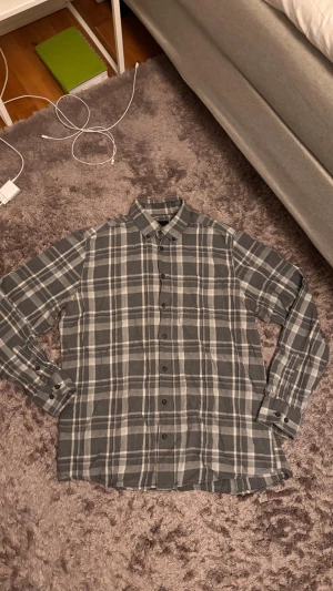 Grå rutig grisch skjorta - Grisch/archive skjorta. Snygg grå skjorta från Dressmann i storlek L med klassiskt rutigt mönster i grått och vitt. Skjortan har långa ärmar, knappar hela vägen och en mjuk bomullskänsla. Perfekt för dig som gillar stilrena och chill plagg. Passar mindre än L