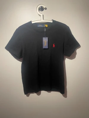 Svart t-shirt från Polo Ralph Lauren - Svart t-shirt från Polo Ralph Lauren med klassisk rund hals och korta ärmar. T-shirten har en röd broderad Polo-logga på bröstet och är gjord i mjuk bomull. Enkel och stilren design som funkar till allt. Märkeslapp kvar på plagget.