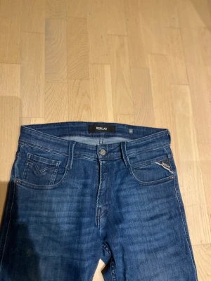 Replay Anbass - Snygga blå jeans från Replay med klassisk femficksdesign och läderpatch bak. Jeansen har smal passform, raka ben och subtila slitningar på framsidan. Materialet är mjukt jeans och midjan är normal. Perfekta för dig som gillar stilrena och moderna jeans.