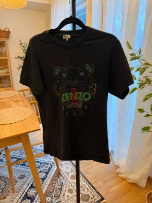 Svart Kenzo t-shirt med tigertryck - Svart t-shirt från Kenzo med ikoniskt tigertryck i färgerna grönt, blått och rött på bröstet. Klassisk rund hals och korta ärmar. Materialet är mjuk bomull som känns skön mot huden. Perfekt för dig som gillar streetstyle och vill sticka ut med ett coolt motiv.