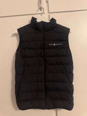 Svart dunväst från Sail Racing - Svart dunväst från Sail Racing med hög krage och dragkedja framtill. Västen har två snedställda fickor med dragkedja och diskret logga på bröstet. Perfekt för lager-på-lager och en clean look.