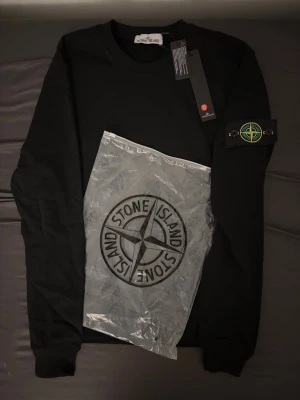 Svart sweatshirt från Stone Island - Svart långärmad sweatshirt från Stone Island med klassisk rund hals och ikonisk patch på vänster ärm. Tröjan har ribbade muddar och är tillverkad i mjuk bomull. Perfekt för dig som gillar streetwear och vill ha en clean look.