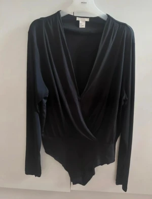 Svart omlott body från H&M - Svart långärmad bodysuit från H&M med omlott-design och djup v-ringning. Passar till både byxa och kjol. Perfekt för att skapa en clean och stilren look