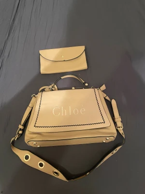 Beige axelväska och plånbok från Chloé - Snygg beige axelväska från Chloé med matchande plånbok. Väskan har ett brett axelband med stora guldfärgade öljetter, handtag med gulddetaljer och broderad logga framtill. Materialet är mjukt skinn och designen är stilren med svarta kanter och dekorativ söm.