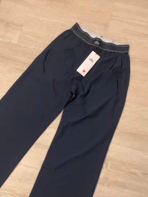 Mörkblå mjukisbyxor från Alo - Snygga mörkblå mjukisbyxor från Alo med bred elastisk midja och diskret logga framtill. Byxorna har en rak och loose passform med mjukt material som är perfekt för chill eller träning. Stilren design med vita detaljer på midjan.