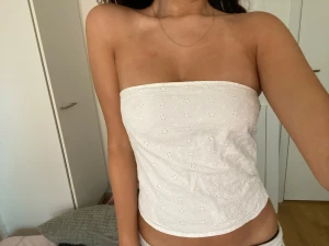 Vit bandeau tubtopp med broderade blommor - Säljer en vit bandeau tubtopp med broderade små blommor. Toppen är axelbandslös och har en lätt croppad passform. Materialet är mjukt bomullstyg med hålbroderi som ger en fräsch och somrig vibe. Perfekt för dig som gillar minimalistisk och söt stil.