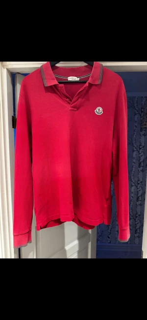 Moncler Pikétröja  - Snygg röd långärmad pikétröja från Moncler med klassisk krage och gråa ränder på kragen. Tröjan har Moncler-logga broderad på bröstet och är gjord i bomull. Använt skick men inga defekter som fläckar eller hål/skador. Storlek L men passar M.