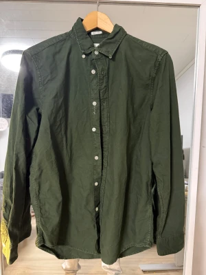 Grön skjorta från East West, S - Snygg mörkgrön skjorta från East West i ekologisk bomull. Klassisk krage, långa ärmar och vita knappar framtill. Skjortan har en clean look utan mönster och passar dig som gillar enkel stil med lite edge.