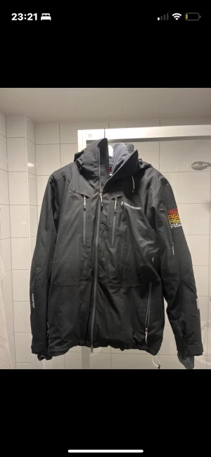 Svart vinterjacka Peak Performance S - Peak Performance vinterjacka i svart med huva, flera dragkedjor och broderad logga på bröstet och ärmen. Jackan är fodrad, har justerbar midja och är tillverkad i slitstark Gore-Tex. Perfekt för kalla dagar och outdoor-aktiviteter.