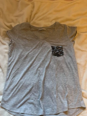 Grå t-shirt med paljettficka  - Grå t-shirt från LCW Casual med v-ringning och korta ärmar. På bröstet finns en liten ficka täckt av svarta och silvriga paljetter storlek M💕
