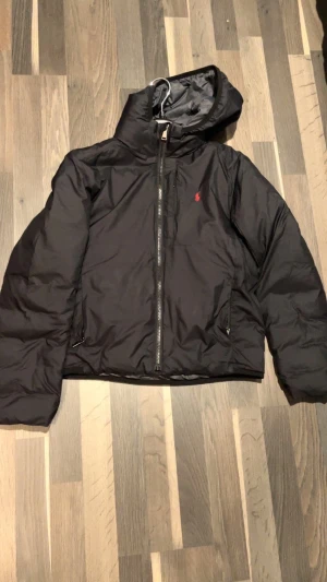 Svart pufferjacka från Polo Ralph Lauren - Svart pufferjacka från Polo Ralph Lauren med huva och dragkedja framtill. Jackan har en röd Polo-logga på bröstet och två fickor med dragkedja. Perfekt för kyliga dagar och har en stilren look som funkar till det mesta. Byter!