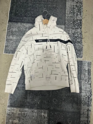 Vit hoodie från BOSS med grafiskt mönster - Säljer en vit hoodie från BOSS med svart grafiskt mönster och en svart linje över bröstet. Tröjan har huva med snörning och en stor magficka. Perfekt för dig som gillar stilrena streetwear-plagg med en twist.