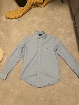 Ljusblå skjorta Polo Ralph Lauren S - Snygg ljusblå skjorta från Polo Ralph Lauren i slim fit. Klassisk design med knappar framtill, lång ärm och ikonisk broderad logga på bröstet. Skjortan är gjord i mjuk bomull och har en stilren krage. Perfekt för dig som gillar clean och tidlös stil. Bara använd 2 gånger i cirka 1 timme